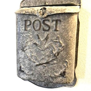 May include: A wall-mounted galvanized metal mailbox with a weathered, mottled gray finish. The front features the word POST embossed in capital letters above a decorative, raised floral or leaf-like pattern. It has a hinged lid at the top.