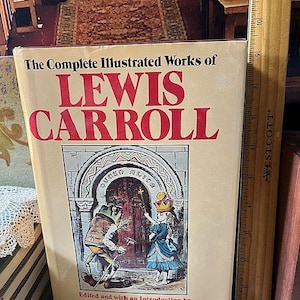 May include: A hardcover book titled "The Complete Illustrated Works of Lewis Carroll." The cover features a red title and an illustration of Alice and a frog-like character. A wooden ruler is placed next to the book.