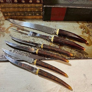 May include: A set of six silver knives with brown and yellow handles. The knives are arranged in a row on a white surface.