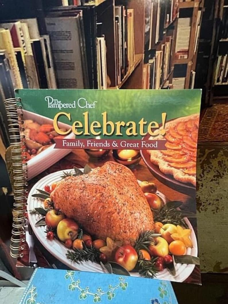 May include: A cookbook titled "The Pampered Chef Celebrate! Family, Friends & Great Food" with a spiral binding. The cover features a roasted turkey breast with apples, cranberries, and rosemary on a white platter.