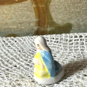May include: A small, porcelain figurine of a kneeling figure with a white head covering, blue robe, and yellow shawl. The figurine is on a white, crocheted surface.