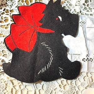 May include: A black felt Scottish Terrier dog-shaped ornament with a large red bow. The dog has white stitched details and a small red eye. The ornament is on a white lace surface.