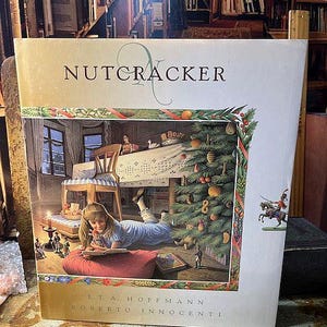 May include: A hardcover book titled "Nutcracker" with a detailed illustration. The cover art depicts a young girl reading, a decorated Christmas tree, and miniature figures. The author's name, E.T.A. Hoffmann, and the illustrator's name, Roberto Innocenti, are also visible.