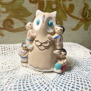 May include: A ceramic owl figurine with a light brown body, white accents, and blue eyes. The owl is surrounded by small figures of children. The figurine is placed on a white, crocheted surface.