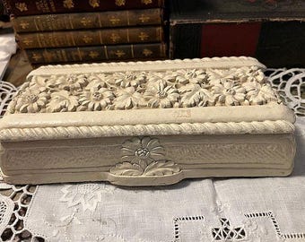 May include: A rectangular off-white decorative box featuring a relief carving of daisy-like flowers and leaves on the lid. The edges of the lid are framed with a rope-style border, and a single flower motif is centered on the front side panel.