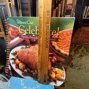 May include: Two cookbooks with the title "The Pampered Chef Celebrate! Family, Friends, Food" on the cover. The cookbooks are open to pages with images of food. A wooden ruler is standing upright between the cookbooks.