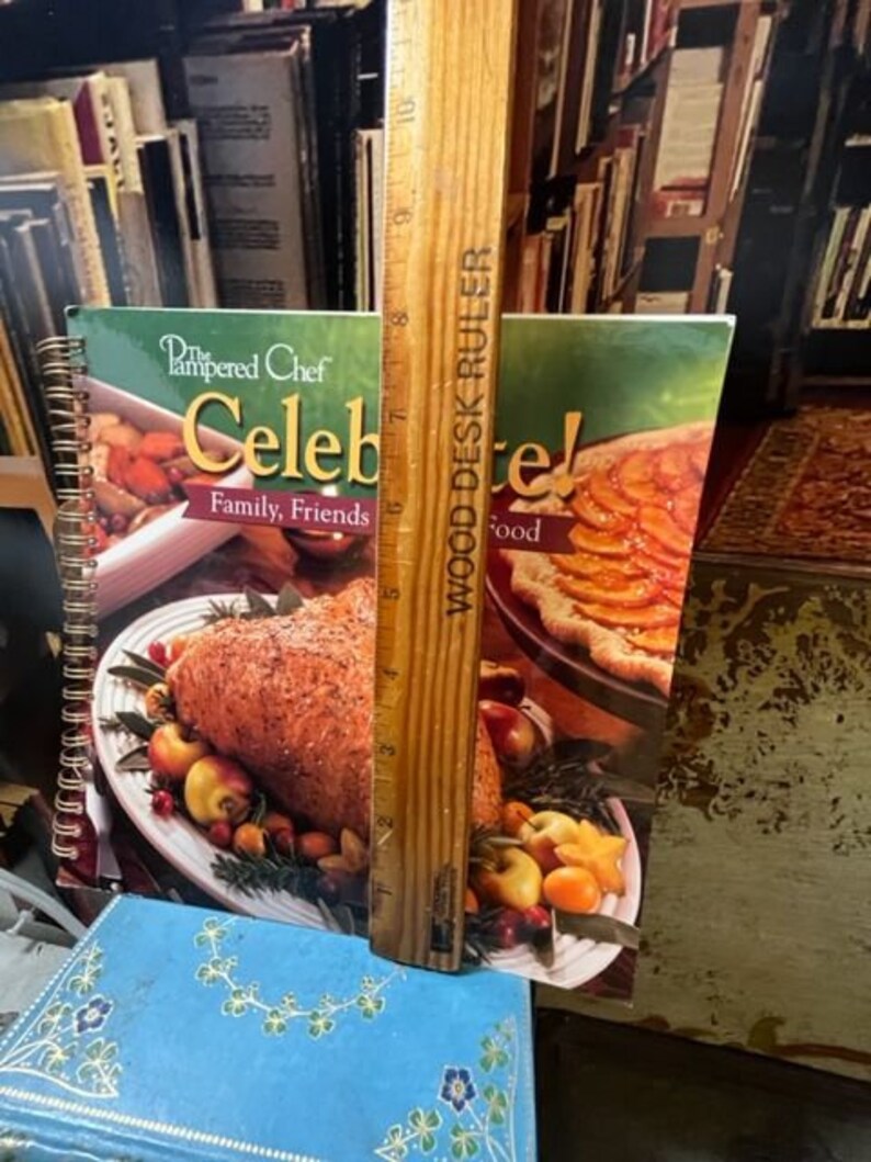 May include: Two cookbooks with the title "The Pampered Chef Celebrate! Family, Friends, Food" on the cover. The cookbooks are open to pages with images of food. A wooden ruler is standing upright between the cookbooks.