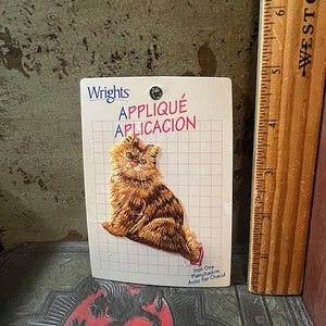 May include: A vintage iron-on appliqué featuring a detailed orange and brown tabby cat. The appliqué is on a grid-patterned card with the text "Wright's APPLIQUE APLICACION". A wooden ruler is next to the card.