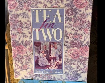 May include: A decorative box with the words "TEA for TWO" in a purple frame, featuring a floral pattern in pink and purple. The box sits atop a blue book with gold floral accents. The box also has the text "Taking Time for Friends".
