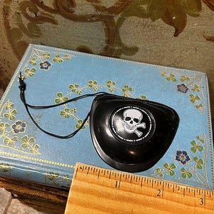 May include: Black pirate eye patch with a white skull and crossbones design. The eye patch is on a blue book with floral and shamrock patterns. A wooden ruler is next to the eye patch.