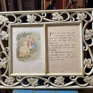 May include: Cream-colored double picture frame with a decorative leaf and scroll design. One side features a vintage illustration of rabbits, the other side has text from a storybook. A charming piece for displaying art or photos.