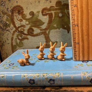 May include: Four small brown plastic bunny figurines on a blue and gold patterned book. The bunnies are arranged in a row, facing forward.