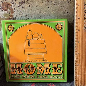 May include: A square, green and orange Snoopy-themed decorative item. The image features Snoopy lying on top of his dog house. The text "HOME IS ON TOP OF A DOG HOUSE" is displayed in a decorative font. The item is next to a wooden ruler.