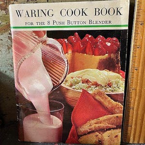 May include: A vintage cookbook titled "WARING COOK BOOK FOR THE 8 PUSH BUTTON BLENDER" with a colorful cover. The cover features images of food, including a strawberry-topped dessert, a dish with a creamy topping, and a glass of pink liquid being poured.