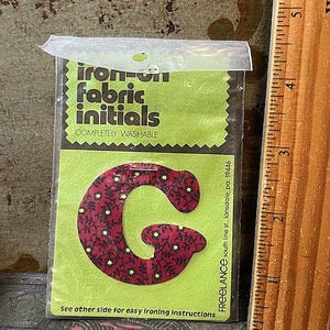 May include: A package of iron-on fabric initials. The package features a lime green background with the words "iron-on fabric initials" in black. A red fabric letter "G" with a floral pattern is visible. The package also includes the text "COMPLETELY WASHABLE".