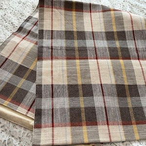 May include: A rectangular fabric runner featuring a plaid pattern in shades of brown, cream, and grey, accented with thin vertical and horizontal stripes in yellow and red. The textile has a visible woven texture and straight, finished edges.