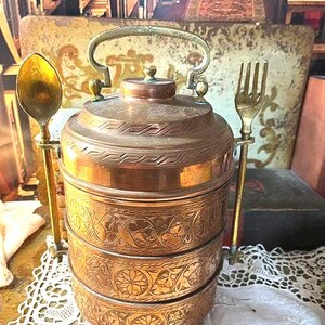 May include: A three-tiered copper tiffin carrier featuring intricate floral engravings on each level. The container has a central handle on the lid and two vertical side attachments, one holding a brass spoon and the other a brass fork.