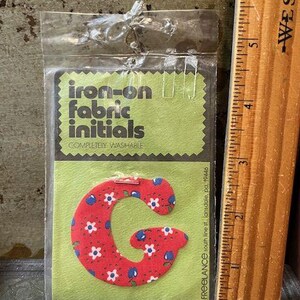May include: A package containing an iron-on fabric initial "G". The initial is red with a floral pattern of white flowers and blue accents. The package also includes the text "Iron-on fabric initials" and a wooden ruler.