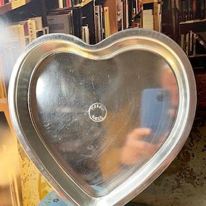 May include: A heart-shaped, silver-colored metal tray. The tray has a smooth, reflective surface and a slightly raised edge. A small, circular emblem is visible in the center. The tray is sitting on a blue surface with a floral pattern.