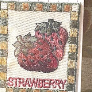 May include: A square embroidered patch featuring two red strawberries with green leaves, centered within a checkered border of brown, tan, and cream squares. The word STRAWBERRY is embroidered in red capital letters at the bottom of the patch.