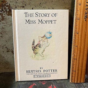 May include: A vintage children's book titled "The Story of Miss Moppet" by Beatrix Potter. The cover features a light brown and white cat holding a blue ball with a white flower on it.