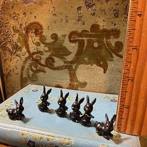 May include: Seven black rabbit figurines lined up on a blue book with a gold spine. The rabbits are all facing forward with their ears up and their tails down. The rabbits are made of a smooth, black material and have red eyes.