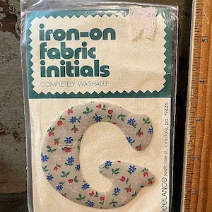 May include: A package of iron-on fabric initials. The package contains a floral patterned letter "G" in beige, red, and blue. The package reads "iron-on fabric initials" and "completely washable".