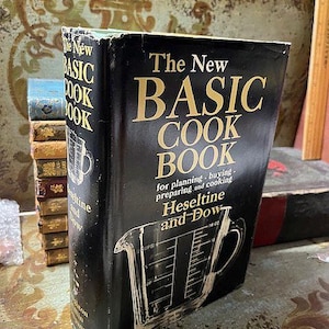 May include: A vintage cookbook titled "The New Basic Cook Book" with a black cover and gold lettering. The cover features a drawing of a measuring cup. The book is slightly worn, suggesting it's a well-used kitchen resource.