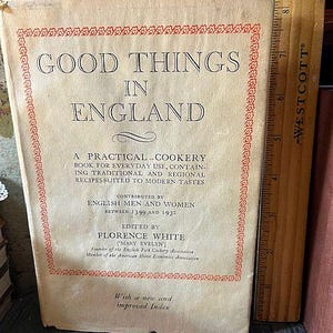 May include: A vintage cookbook titled "Good Things in England" with a cream-colored cover and red decorative border. The book includes text about traditional recipes and is edited by Florence White. A wooden ruler is placed next to the book.