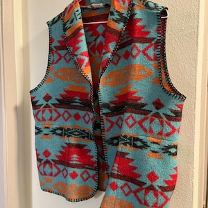 May include: A turquoise vest with a Southwestern pattern in red, orange, and black. The vest is sleeveless with a collar and is hanging on a white hanger. The edges are finished with black stitching.