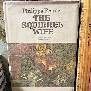 May include: A hardcover book titled "The Squirrel Wife" by Philippa Pearce, with illustrations by Derek Collard. The cover features a squirrel amidst foliage in shades of brown, green, and orange. A wooden ruler is positioned next to the book.