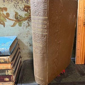 May include: A vintage book titled "About Papers Four Georges and English Humorists" with embossed floral patterns on the spine and cover. The book is brown and stands upright, showcasing its aged appearance. A wooden ruler is visible in the background.