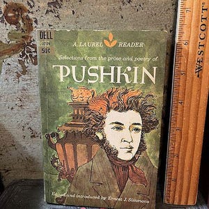 May include: Vintage paperback book titled "Pushkin" from the Laurel Reader series. The cover features a portrait of a man with reddish-brown hair and a brown jacket. A wooden ruler is on the right side of the book.