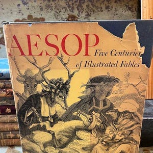 May include: A vintage book titled "AESOP Five Centuries of Illustrated Fables." The cover features an illustration of anthropomorphic animals in a sepia tone. The book's cover is aged with wear and tear.