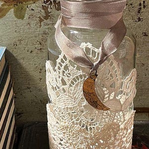 May include: A clear glass jar wrapped with cream-colored lace and a taupe ribbon tied around the neck. A bronze crescent moon charm hangs from the ribbon. The jar is a decorative item.