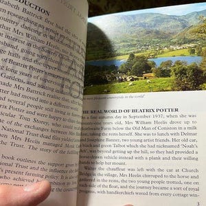 May include: Open book with text and a color image of a landscape with a lake, mountains, and trees. The text discusses Beatrix Potter and her life in the countryside.