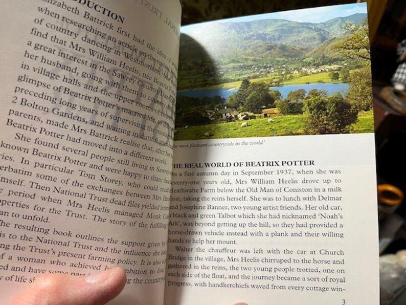 May include: Open book with text and a color image of a landscape with a lake, mountains, and trees. The text discusses Beatrix Potter and her life in the countryside.