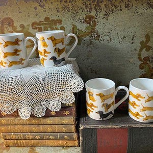 May include: White ceramic mugs with a repeating pattern of dachshund dogs in various poses and colors. The mugs have a curved handle and are displayed on a stack of vintage books and a lace doily.