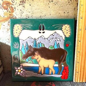 May include: A square ceramic tile featuring a dark brown adult moose and a light tan calf standing near a blue river. The background shows mountains and pine trees. A wooden ruler on the right indicates the tile is approximately 6 inches tall.