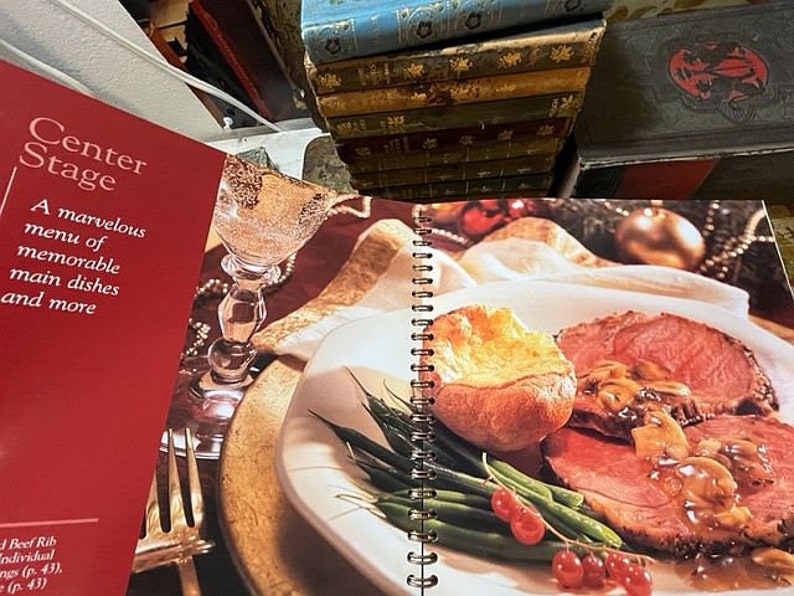 May include: A cookbook titled "Center Stage" with a red cover and the text "A marvelous menu of memorable main dishes and more". The book is open to a page showing a plate of roast beef with Yorkshire pudding, green beans, and red currants.