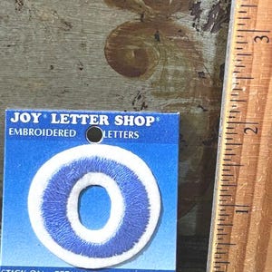 May include: A blue and white embroidered letter O patch on a blue Joy Letter Shop card. The patch is positioned next to a wooden ruler showing a height of approximately 1.5 inches for the letter. The packaging text reads Stick On, Permanent, Iron On.