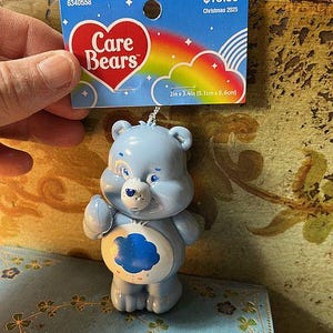 May include: A light blue Care Bears ornament with a cloud design on its belly. The ornament is 2 inches by 3.4 inches. A tag with the Care Bears logo and the text "Christmas 2025" is attached.