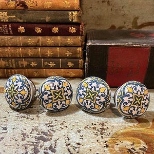 May include: Four decorative cabinet knobs with a cream base, featuring a blue and yellow floral pattern. The knobs are round and sit on a distressed wooden surface, with a stack of antique books in the background.