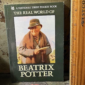 May include: A National Trust Pocket Book titled "The Real World of Beatrix Potter." The cover features a portrait of Beatrix Potter in a hat and coat, holding a paper. The book is green with white text.