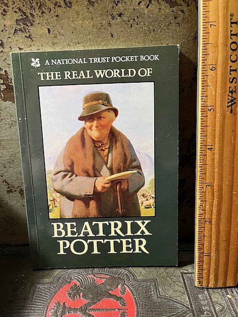May include: A National Trust Pocket Book titled "The Real World of Beatrix Potter." The cover features a portrait of Beatrix Potter in a hat and coat, holding a paper. The book is green with white text.