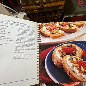 May include: A cookbook page with a recipe for Warm Olive Bruschetta. The recipe includes ingredients like French bread, olive oil, celery, olives, garlic, Italian salad dressing, and cream cheese spread. The page also includes two photos of the finished dish, one with a blue plate and the other with a white plate.