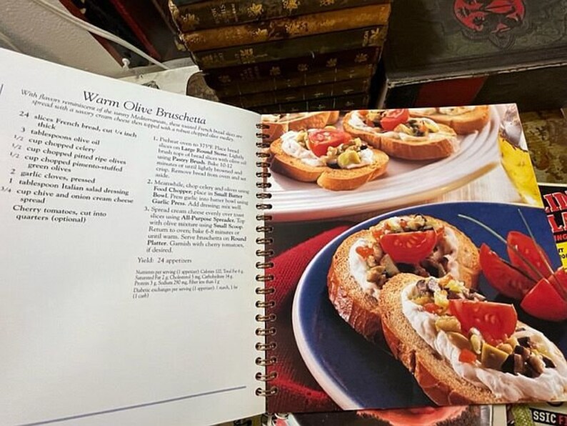 May include: A cookbook page with a recipe for Warm Olive Bruschetta. The recipe includes ingredients like French bread, olive oil, celery, olives, garlic, Italian salad dressing, and cream cheese spread. The page also includes two photos of the finished dish, one with a blue plate and the other with a white plate.