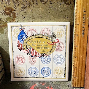May include: A square ceramic tile featuring the seals of the original thirteen states. The tile has a white background with a gold border. The tile includes an American flag and the text "SEALS of The Original Thirteen States".