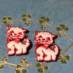 May include: Two embroidered patches of white kittens with red accents, including bows and facial details. The patches are set against a blue background with gold and green floral embellishments.