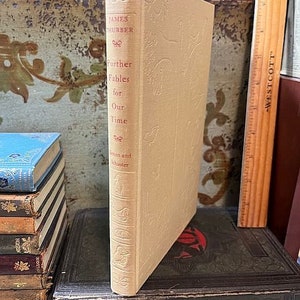 May include: A beige book with the title "Further Fables for Our Time" by James Thurber. The book is on a shelf with other books.